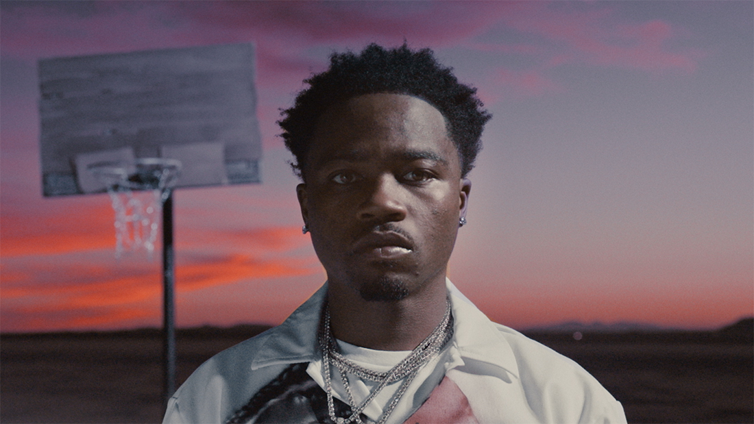 Roddy Ricch stands in front of basketball hoop and pink sky. 